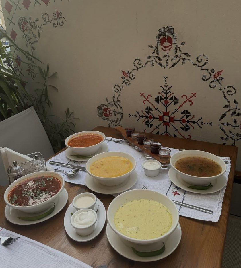 Five bowls of soup with sour cream and green chillies on a wooden table under the painted cross-stitch wall