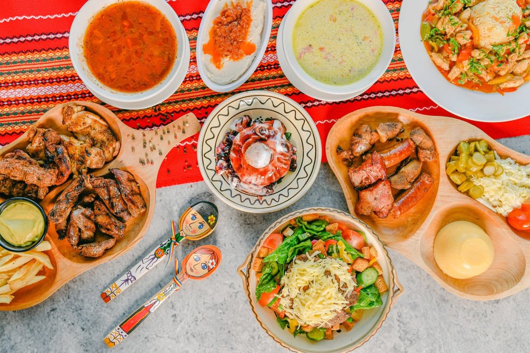 A spread of Romanian dishes on a red woven runner: two soups, grilled meats and sausages on wooden boards, polenta, pickles and a salad