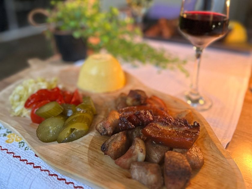 Pork pieces and sausages on a wooden board with pickles, tomato and a dome of polenta, a glass of red wine behind